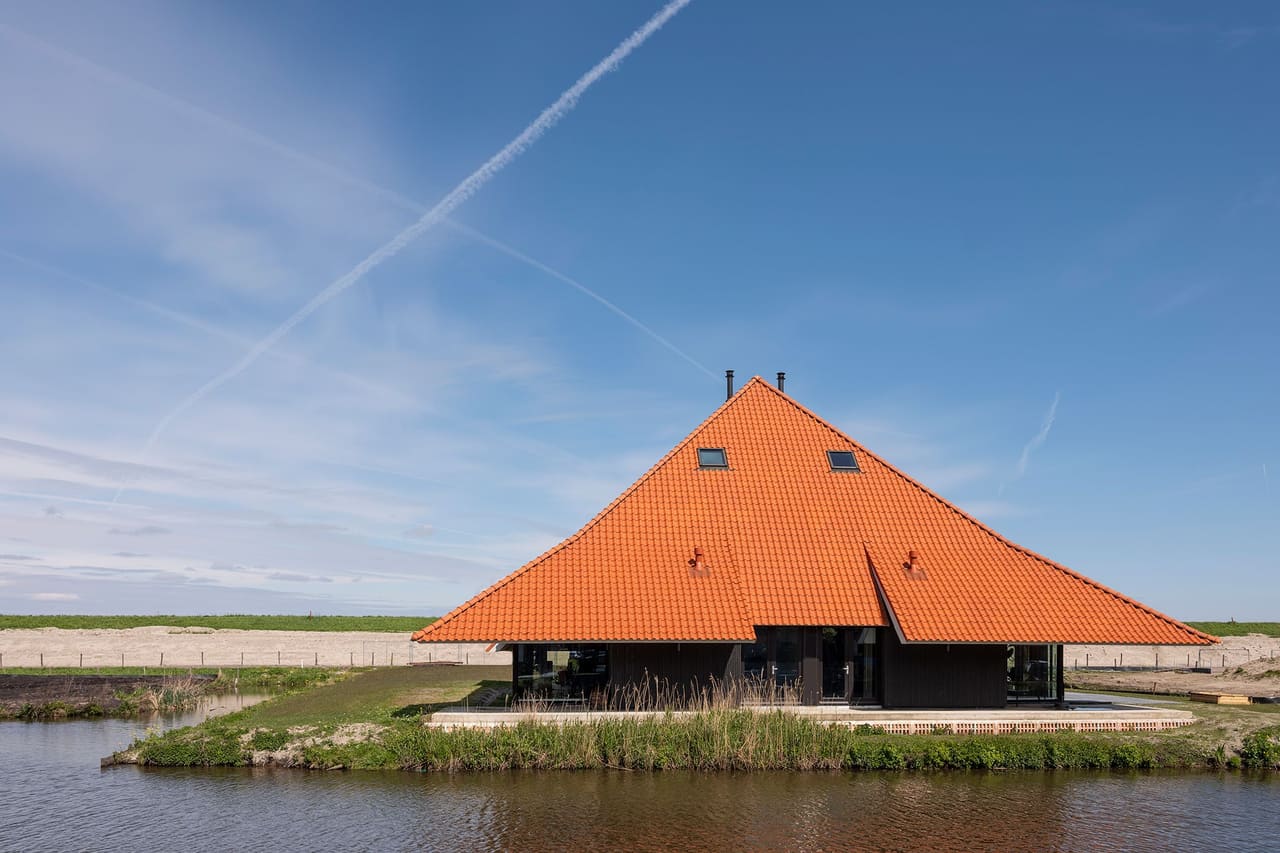 Stolp Zeedijk: een nieuwe stolpboerderij met oranjebruin piramidedak aan het water, onder een blauwe lucht