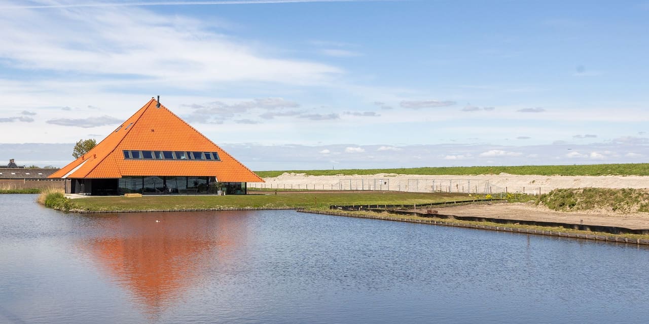 Stolp Zeedijk: de stolpboerderij met oranjebruin piramidedak spiegelt zich in het water, met de dijk aan de horizon